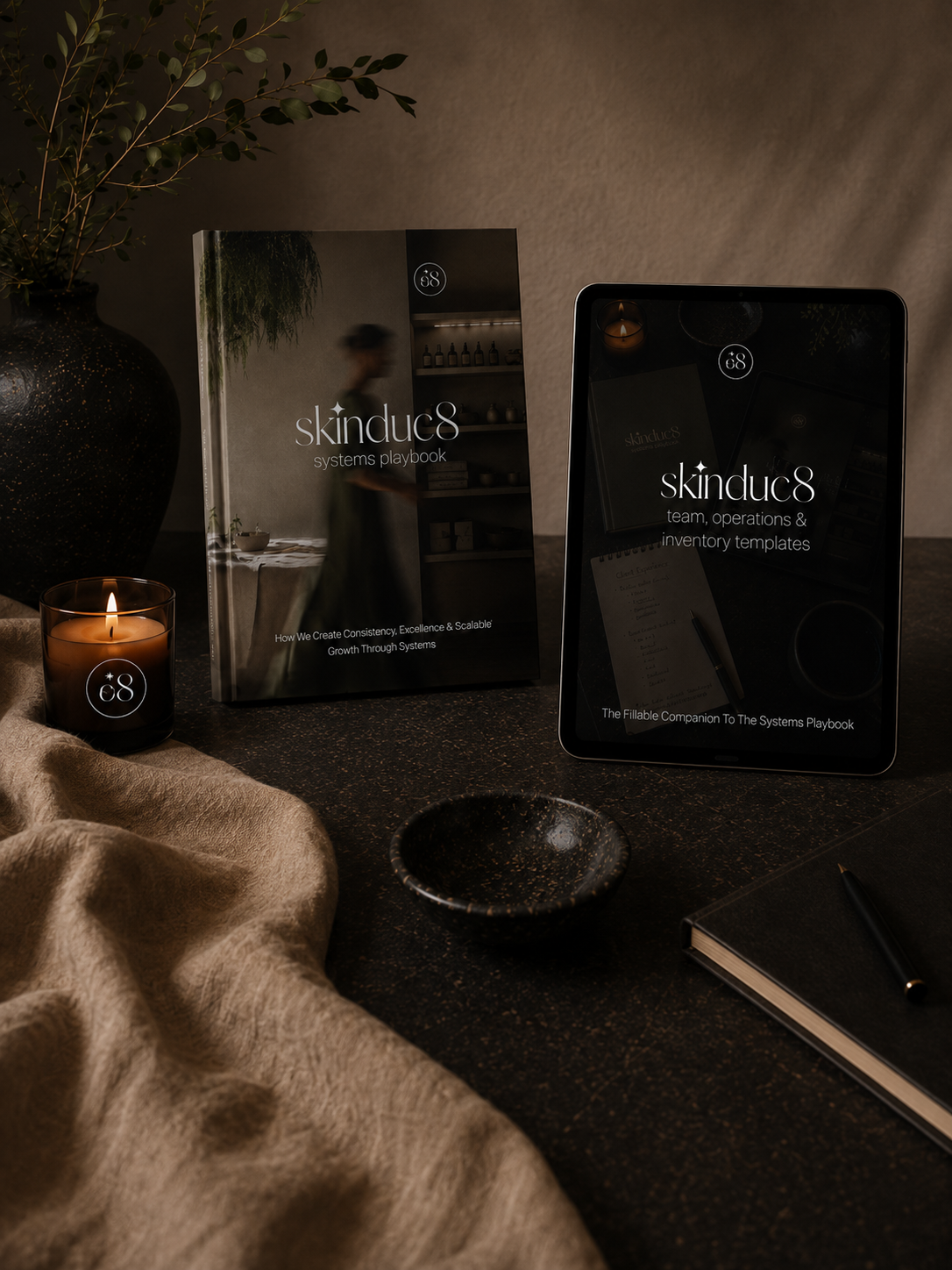 Skinduc8 Systems Playbook hardcover book styled beside an iPad displaying the Team, Operations & Inventory Templates cover, with a lit candle, ceramic bowl, linen throw and eucalyptus branch on a dark textured surface."